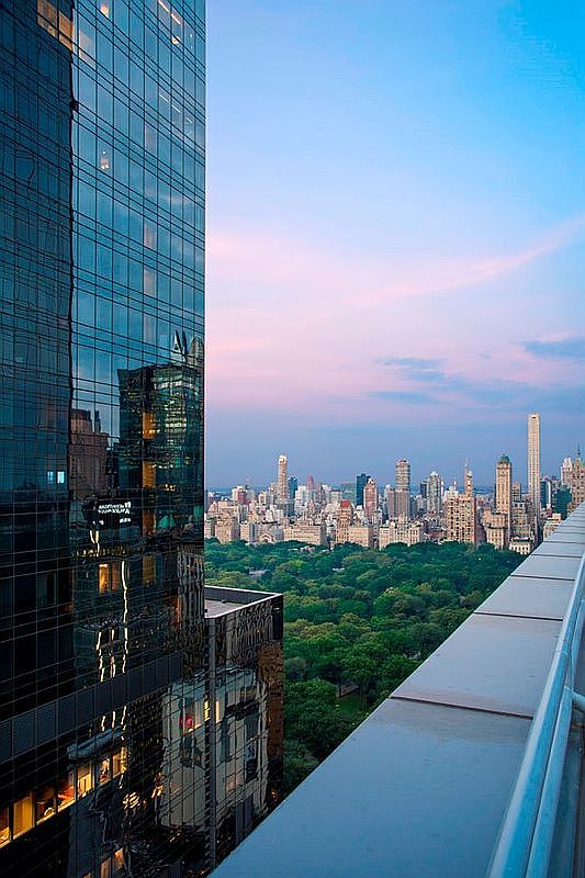 This high-altitude perspective captures a stunning, panoramic view of Central Park and the New York City skyline from a sleek, modern balcony. The foreground features the reflective glass facade of a skyscraper, creating a dramatic contrast between the architectural structure and the lush greenery of the park below. The scene is bathed in the soft, golden light of dusk, emphasizing the luxury and exclusivity of this urban vantage point.