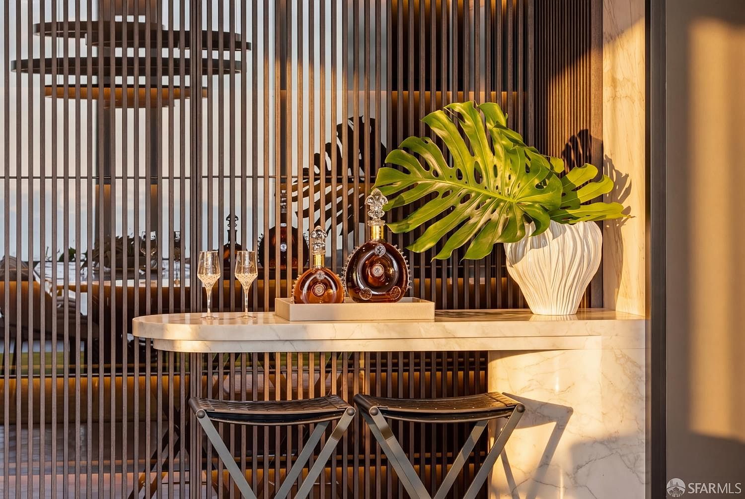 This sophisticated bar area features a sleek, curved stone countertop supported by a matching base, paired with two modern folding stools. Behind the counter, a vertical slat screen creates a textured backdrop, while a large, vibrant monstera leaf in a white sculptural vase adds a touch of organic elegance. Two crystal decanters and matching glasses sit on a tray, bathed in warm, golden light that highlights the refined materials and contemporary design.