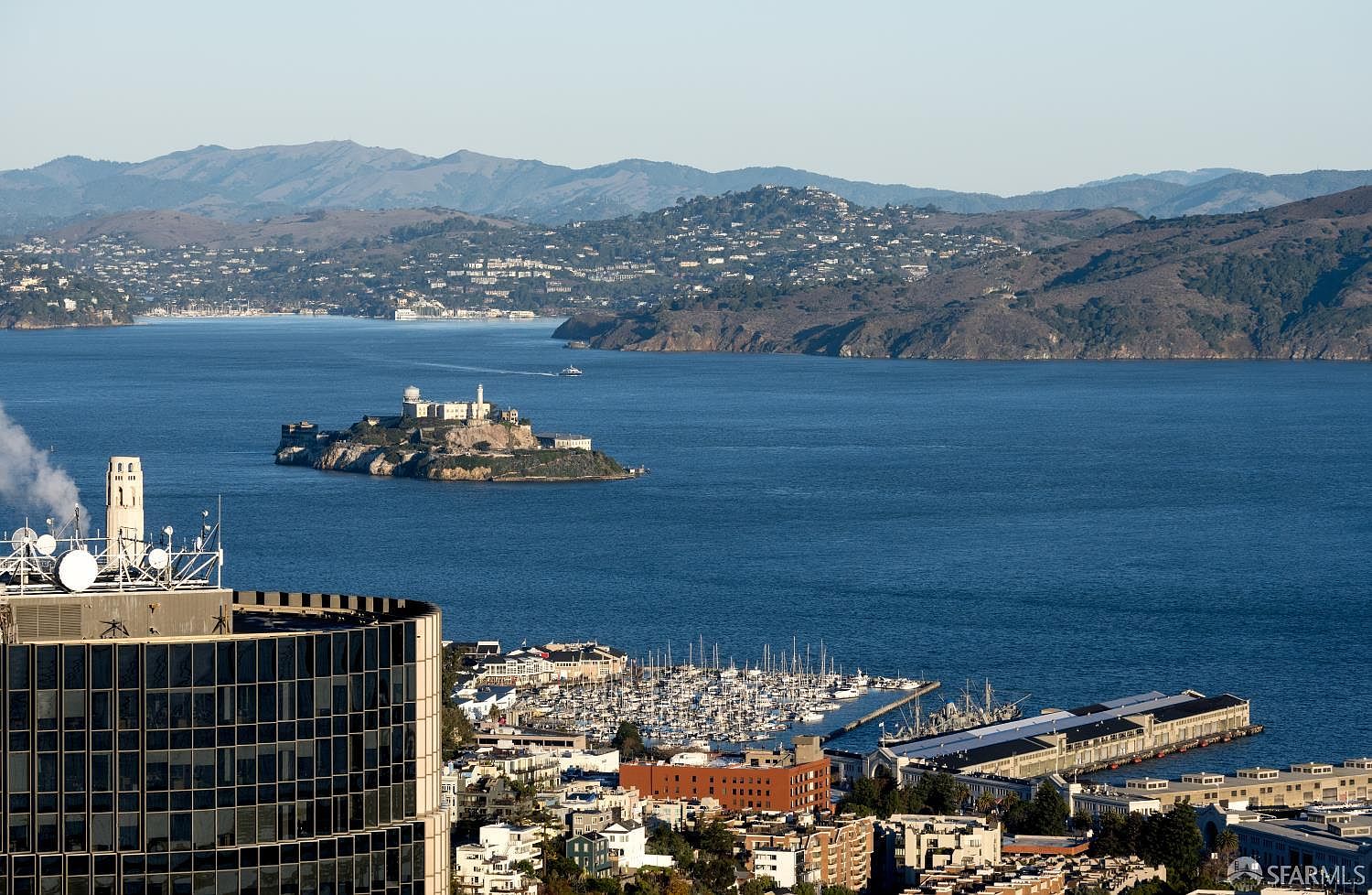 This high-angle aerial view captures a stunning panoramic perspective of the San Francisco Bay, featuring Alcatraz Island prominently in the center. In the foreground, a modern high-rise building with a curved glass facade stands beside a bustling marina filled with numerous boats and a large pier structure. The background showcases the rolling hills of the Marin Headlands under a clear, bright sky, emphasizing the prime coastal location and scenic urban-maritime lifestyle.
