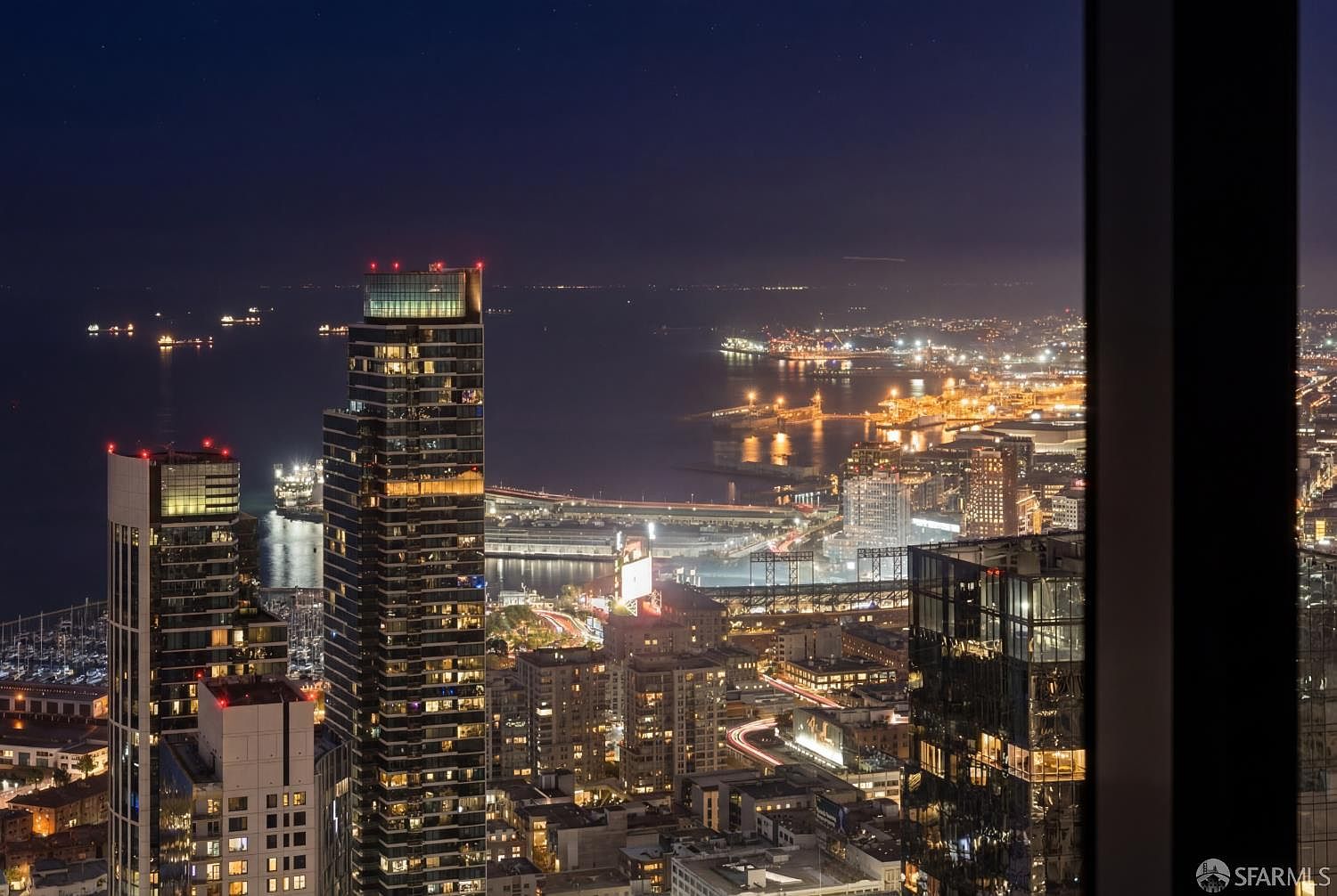 This high-altitude, nighttime aerial view captures a stunning urban skyline from a vantage point within a high-rise building. The scene features illuminated skyscrapers, a bustling harbor with docked boats, and glowing city streets that create a vibrant, cinematic atmosphere. The perspective emphasizes the luxury and height of the property, showcasing an expansive, panoramic cityscape under a dark, starry sky.