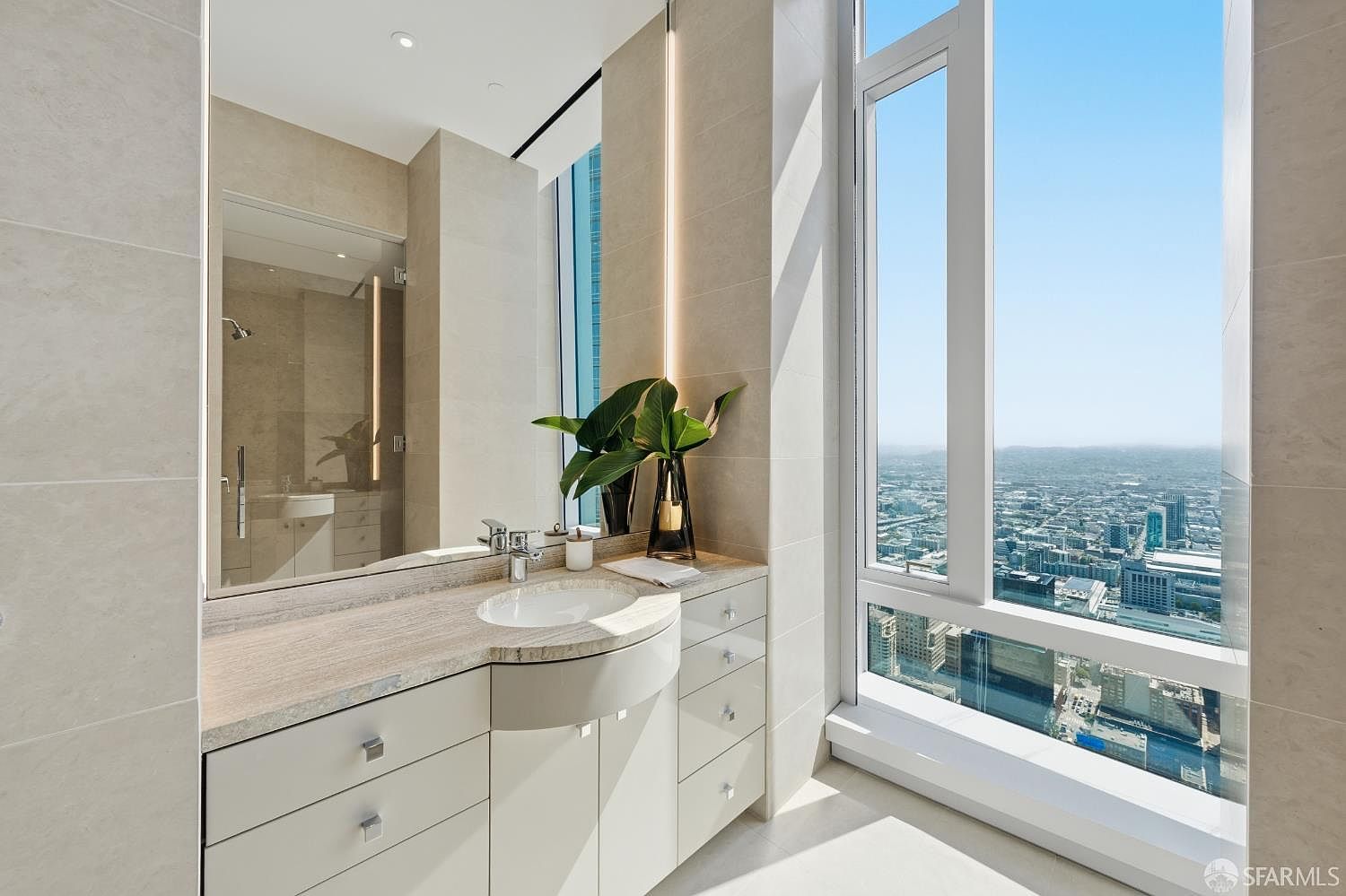This modern bathroom features a sleek vanity with light-colored cabinetry and a stone countertop, complemented by a large mirror reflecting the shower area. A prominent floor-to-ceiling window provides an expansive, unobstructed view of the city skyline, bathing the space in natural light. The minimalist design is accented by a decorative vase with greenery, creating a sophisticated and airy atmosphere.