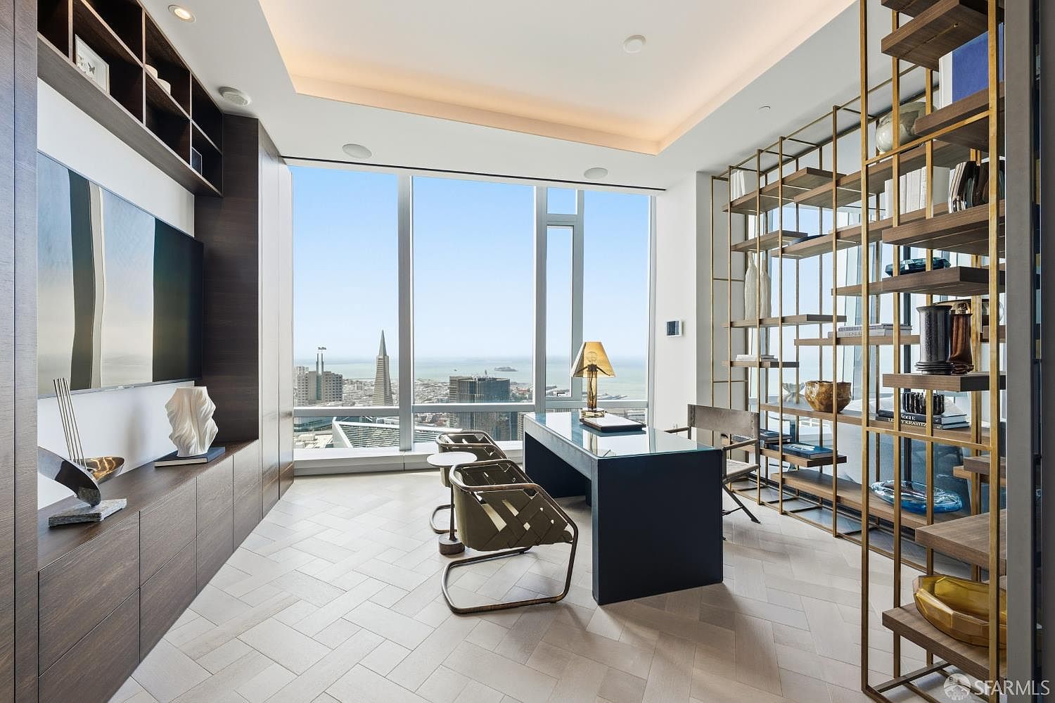 This sophisticated home office features a sleek, dark desk positioned to face floor-to-ceiling windows that offer a panoramic view of the city skyline and bay. The room is accented by a tall, open-frame metal shelving unit and custom dark wood cabinetry, all set against light-toned herringbone flooring. The space exudes a professional and modern atmosphere, perfectly suited for high-end urban living.