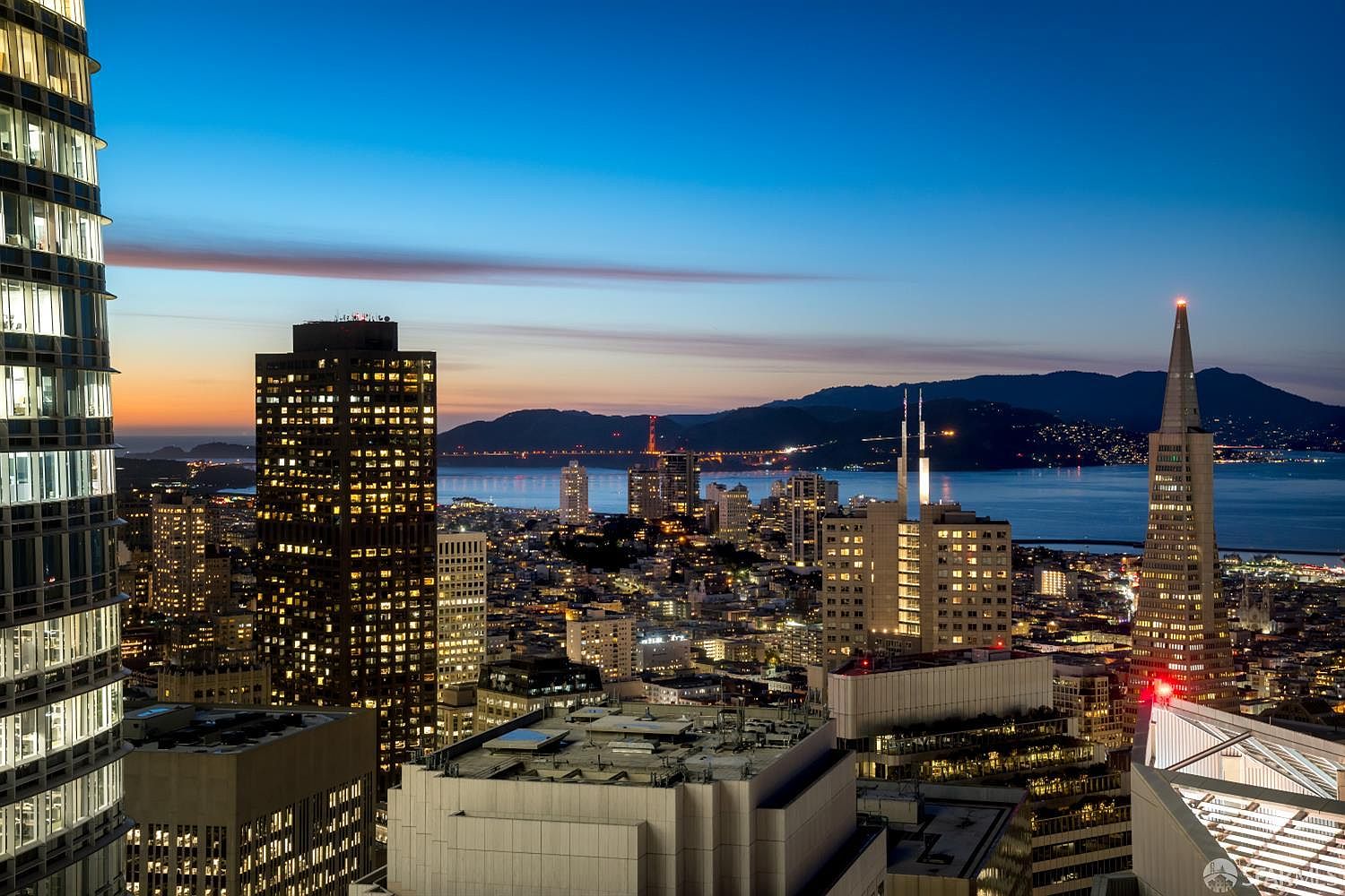 This high-altitude aerial view captures the San Francisco skyline at dusk, showcasing iconic landmarks like the Transamerica Pyramid and the distant Golden Gate Bridge. The perspective is cinematic, looking out over a dense urban landscape illuminated by city lights against a vibrant twilight sky. The foreground features the reflective glass facade of a modern skyscraper, emphasizing the property's prime location and elevated vantage point.