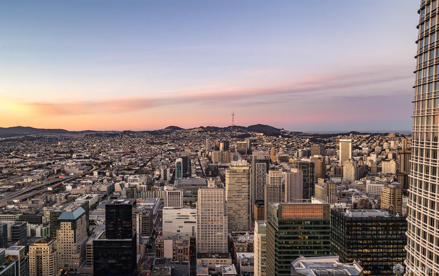 This high-altitude aerial view captures a sprawling urban cityscape at sunset, showcasing a dense grid of skyscrapers and residential buildings. The perspective highlights the dramatic scale of the city, with the golden hour light casting long shadows and painting the sky in soft hues of pink and orange. A prominent, modern high-rise structure dominates the right foreground, framing the expansive metropolitan vista that stretches toward distant, rolling hills.