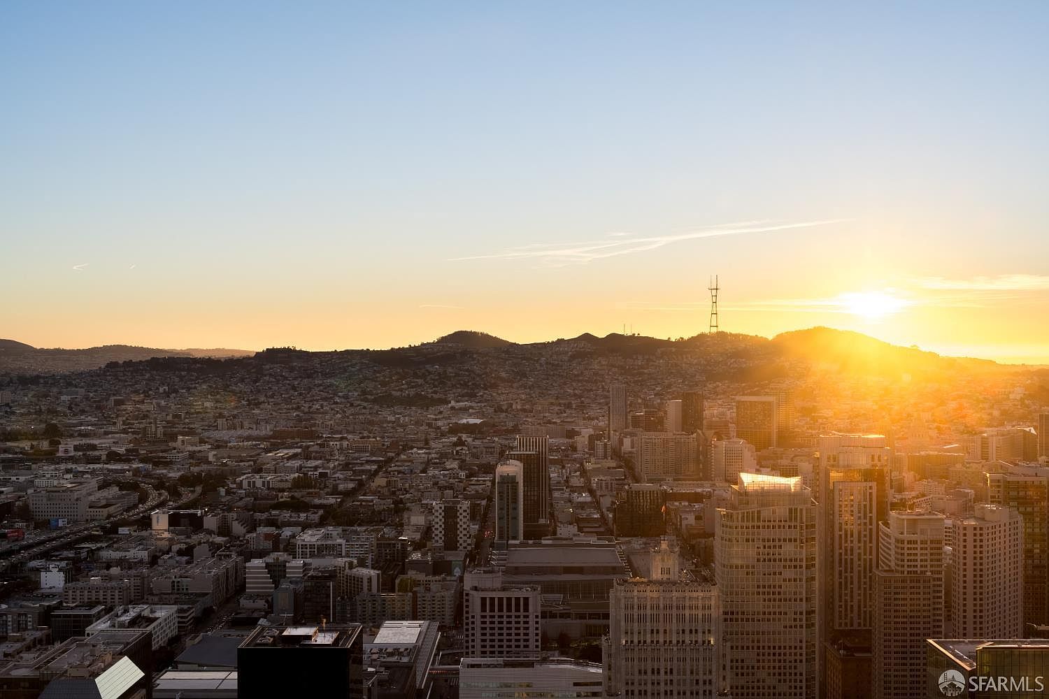This high-angle aerial view captures a sprawling urban cityscape bathed in the warm, golden light of a sunset. The dense grid of buildings stretches toward distant, rolling hills, with a prominent radio tower silhouetted against the bright horizon. The perspective offers a cinematic sense of scale, highlighting the vastness of the metropolitan area during the golden hour.