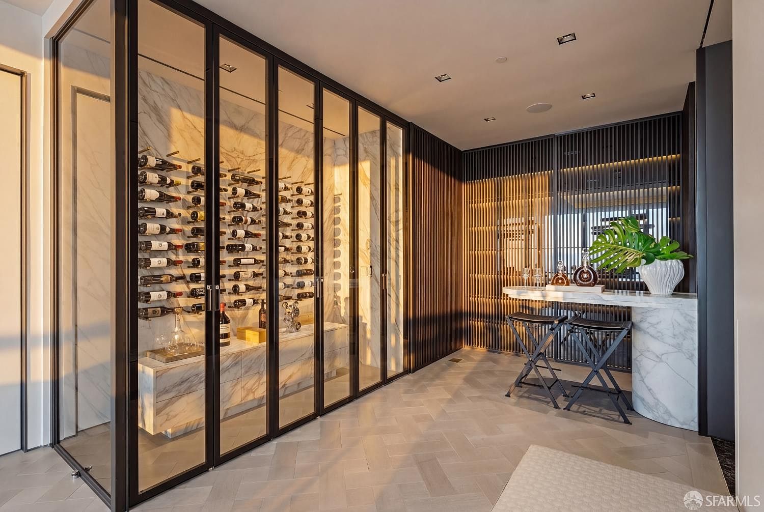 This sophisticated wine cellar features a glass-enclosed display area with stone-lined walls and custom racking for wine storage. Adjacent to the cellar is a sleek bar area with a stone-topped counter, two modern stools, and a decorative plant, set against a backdrop of dark, vertical wood slats. The space is finished with light-toned herringbone flooring, creating a luxurious and modern atmosphere perfect for entertaining.
