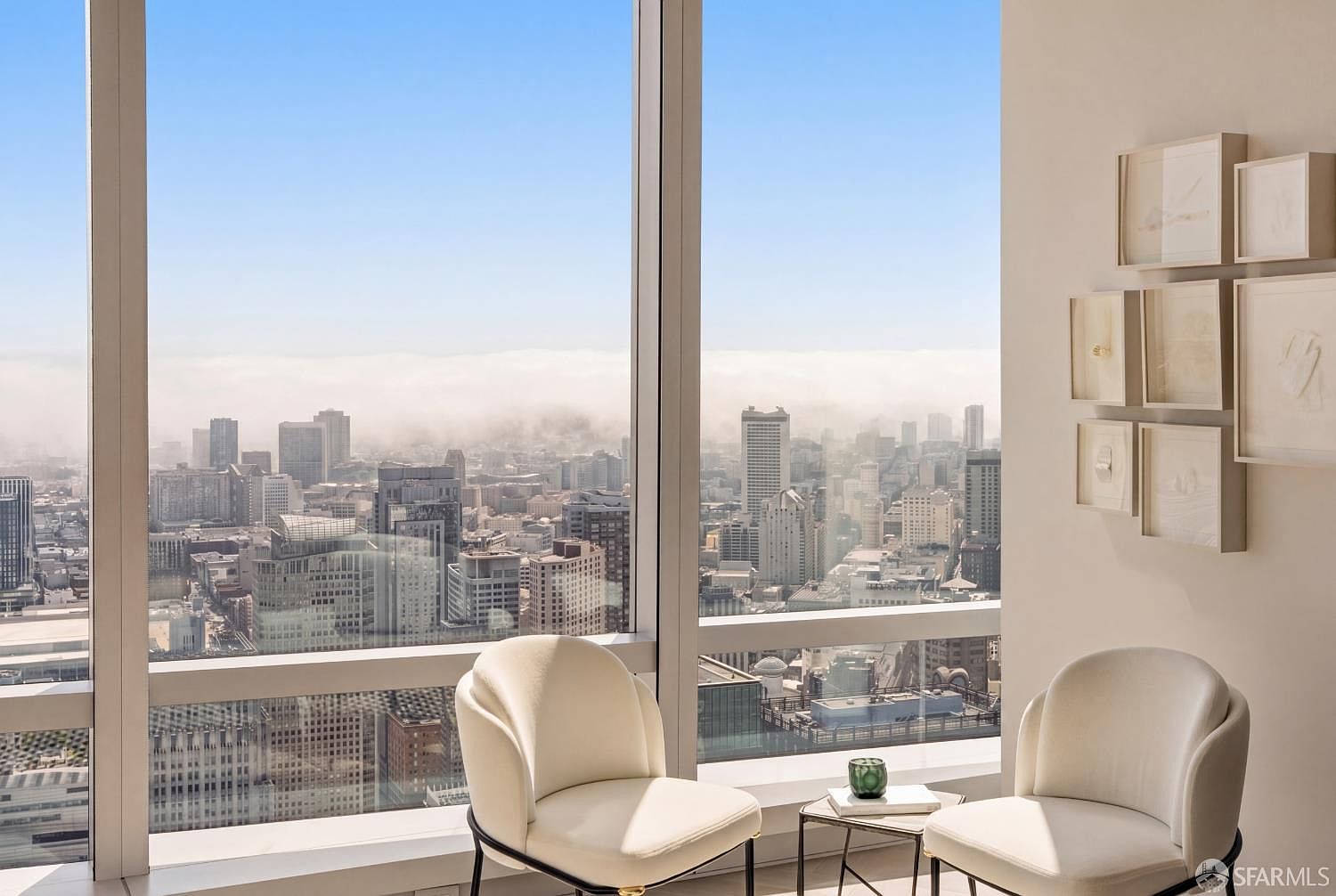 This high-rise living space features expansive floor-to-ceiling windows that frame a panoramic view of the city skyline and a layer of low-hanging clouds. Two modern, cream-colored armchairs are positioned near a small side table, creating an intimate seating area focused on the vista. A gallery wall of framed artwork adds a sophisticated touch to the neutral-toned interior, emphasizing the luxury and height of the residence.