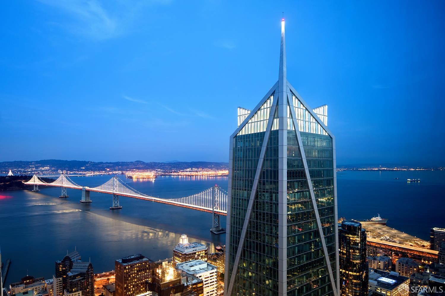 This high-altitude aerial view captures the striking, modern architecture of a prominent skyscraper in San Francisco at twilight. The building features a distinctive, angular glass facade with a prominent spire, set against the backdrop of the illuminated Bay Bridge and the city skyline. The composition emphasizes the building's height and its commanding position overlooking the bay, creating a sophisticated and urban atmosphere.