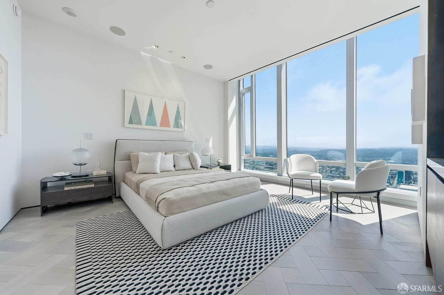 This modern primary bedroom features a minimalist aesthetic with light-toned herringbone flooring and a patterned area rug. A plush bed is centered in the room, flanked by sleek nightstands, while floor-to-ceiling windows provide expansive, unobstructed views of the city skyline. The space is bright and airy, characterized by clean lines and a sophisticated, high-rise urban atmosphere.