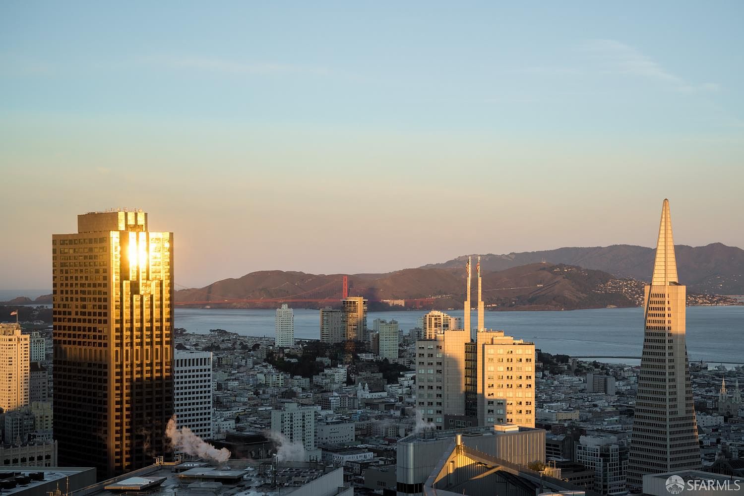 This high-angle aerial view captures a stunning sunset over the San Francisco skyline, featuring the iconic Transamerica Pyramid and the Golden Gate Bridge in the distance. The warm, golden light reflects off the glass facades of the surrounding skyscrapers, creating a dramatic and sophisticated urban atmosphere. The perspective provides a comprehensive look at the dense city architecture set against the backdrop of the bay and rolling hills.