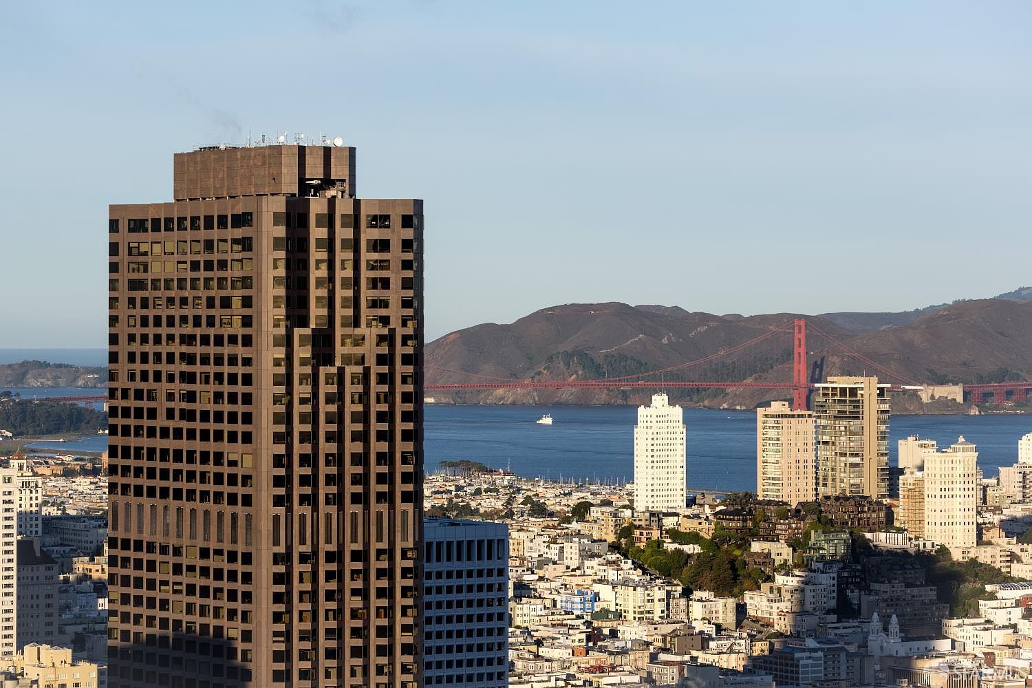 This high-angle aerial view captures a dense urban cityscape, dominated by a prominent, dark-toned skyscraper in the foreground. Beyond the city grid, the iconic Golden Gate Bridge spans the bay, set against a backdrop of rolling hills under a clear sky. The perspective offers a sweeping look at the architectural density and the scenic coastal geography of the area.