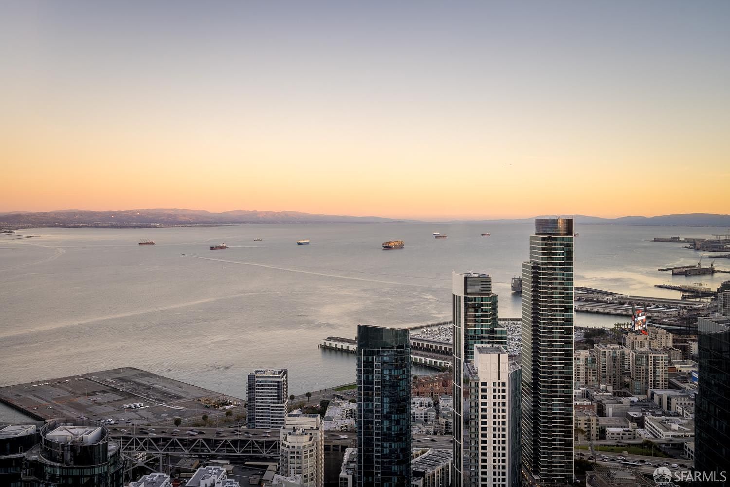 This high-altitude aerial view captures a stunning sunset over the San Francisco Bay, showcasing a dense urban skyline dominated by modern glass skyscrapers. The perspective highlights the juxtaposition of the bustling city architecture against the expansive, calm waters dotted with cargo ships. The golden hour light casts a warm, serene glow over the entire cityscape, emphasizing the prime waterfront location.