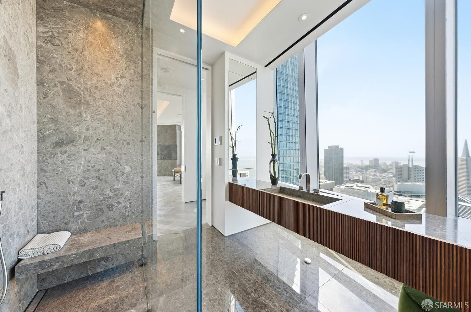 This luxurious primary bathroom features a sleek, modern design with a glass-enclosed shower area containing a built-in stone bench. A long, floating vanity with a slatted wood base and a stone sink sits against expansive windows that provide a panoramic view of the city skyline. The space is finished with polished stone flooring and walls, creating a sophisticated and airy atmosphere.