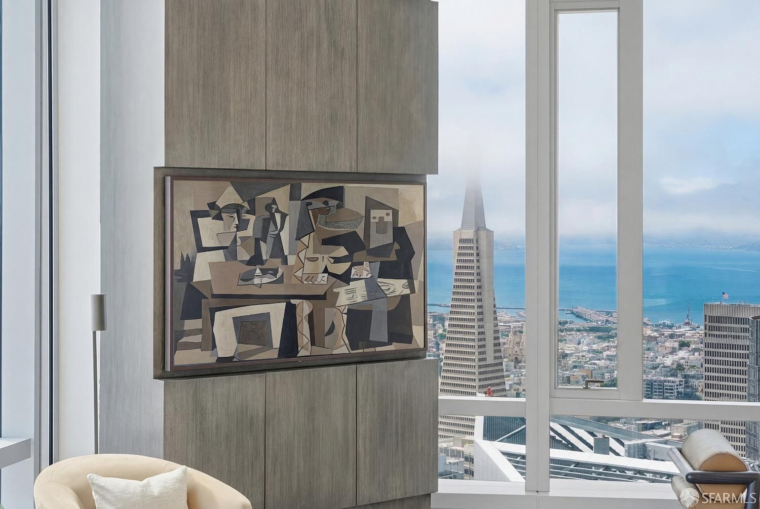 This interior view captures a sophisticated living space featuring a custom wood-paneled wall unit that houses a large, abstract cubist-style painting. To the right, expansive floor-to-ceiling windows provide a dramatic, elevated perspective of the San Francisco skyline, prominently showcasing the Transamerica Pyramid. The composition balances modern architectural millwork with a high-rise urban aesthetic, creating a refined and luxurious atmosphere.