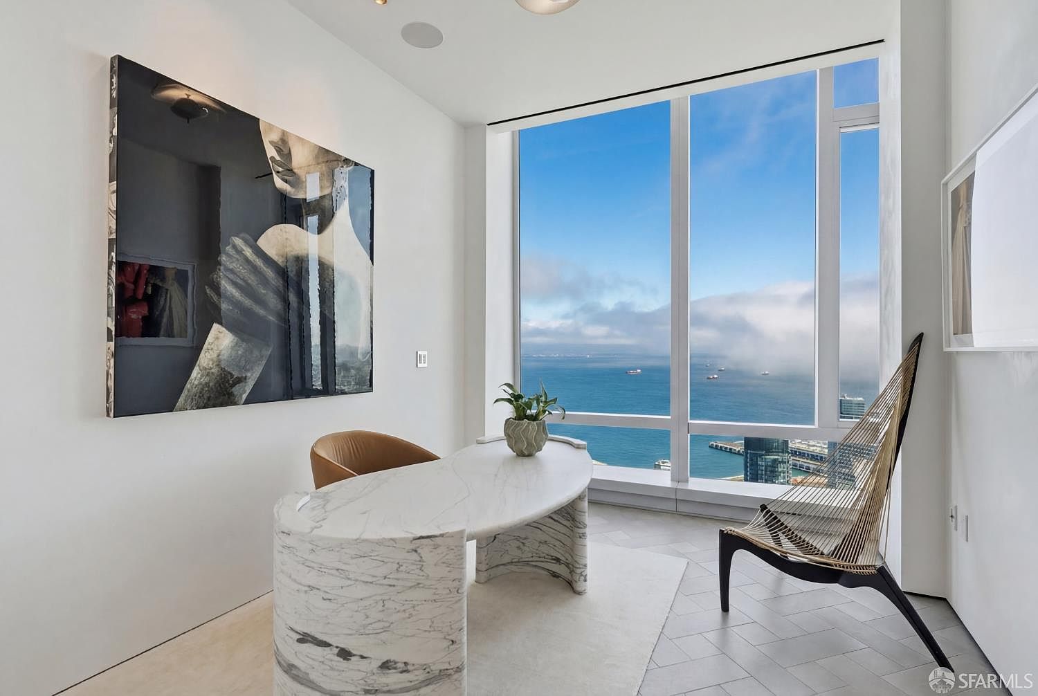 This sophisticated home office features a striking curved stone desk as its centerpiece, positioned to take full advantage of the expansive, floor-to-ceiling windows overlooking a coastal cityscape. A large, bold piece of abstract art hangs on the white wall, providing a dramatic contrast to the minimalist aesthetic and light-toned flooring. The room is bathed in natural light, creating a serene and professional atmosphere perfect for focused work.