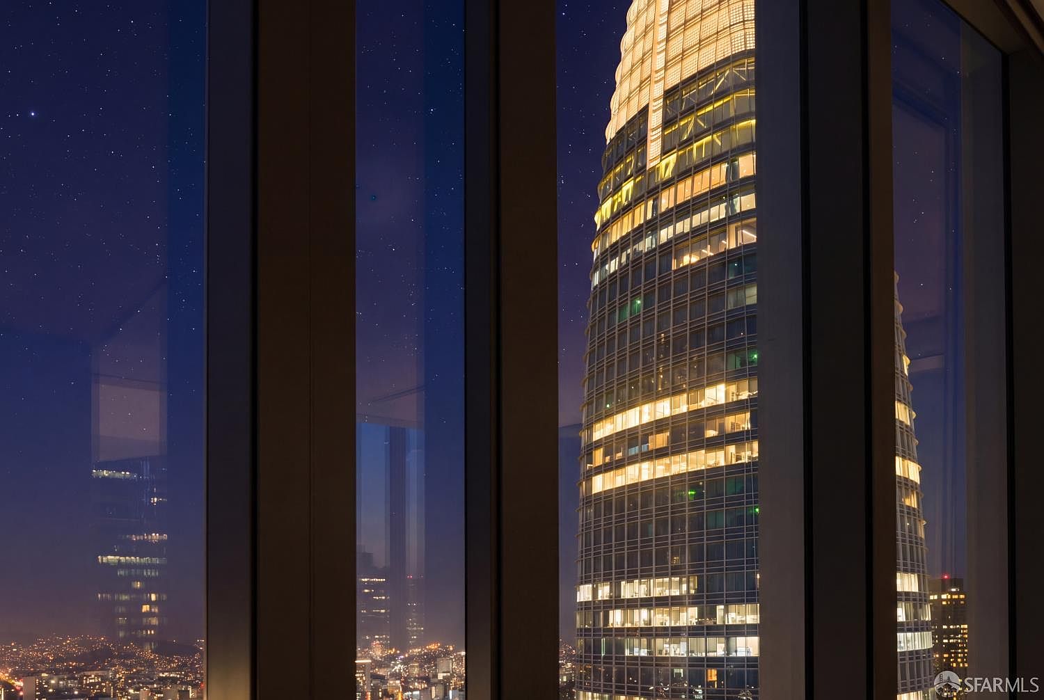 This image captures a dramatic night view from inside a high-rise residence, looking out through floor-to-ceiling windows at the illuminated Salesforce Tower in San Francisco. The perspective is framed by dark vertical window mullions, creating a cinematic contrast between the interior darkness and the glowing, grid-like facade of the skyscraper. The scene evokes a sophisticated, urban atmosphere, highlighting the property's prime location and expansive city views.