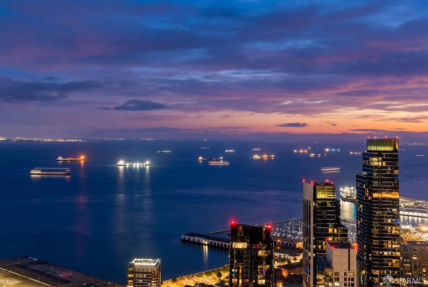 This high-altitude aerial view captures a stunning twilight cityscape overlooking a vast bay filled with cargo ships. The foreground features several modern high-rise buildings with illuminated windows, while the horizon glows with the vibrant colors of a sunset. The perspective emphasizes the urban density and the expansive waterfront setting, creating a sophisticated and dramatic atmosphere.