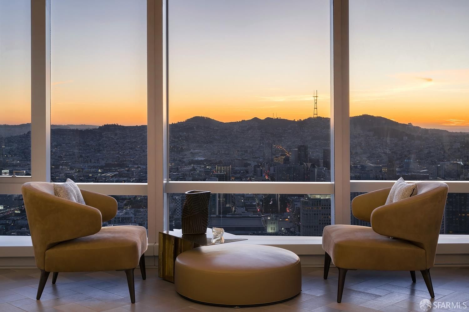 This sophisticated living space features two plush, tan-colored armchairs positioned symmetrically around a low, circular ottoman and a small side table. The room is defined by expansive floor-to-ceiling windows that frame a breathtaking panoramic sunset view of the city skyline and distant hills. The minimalist furniture arrangement emphasizes the dramatic urban vista, creating a serene and luxurious atmosphere.