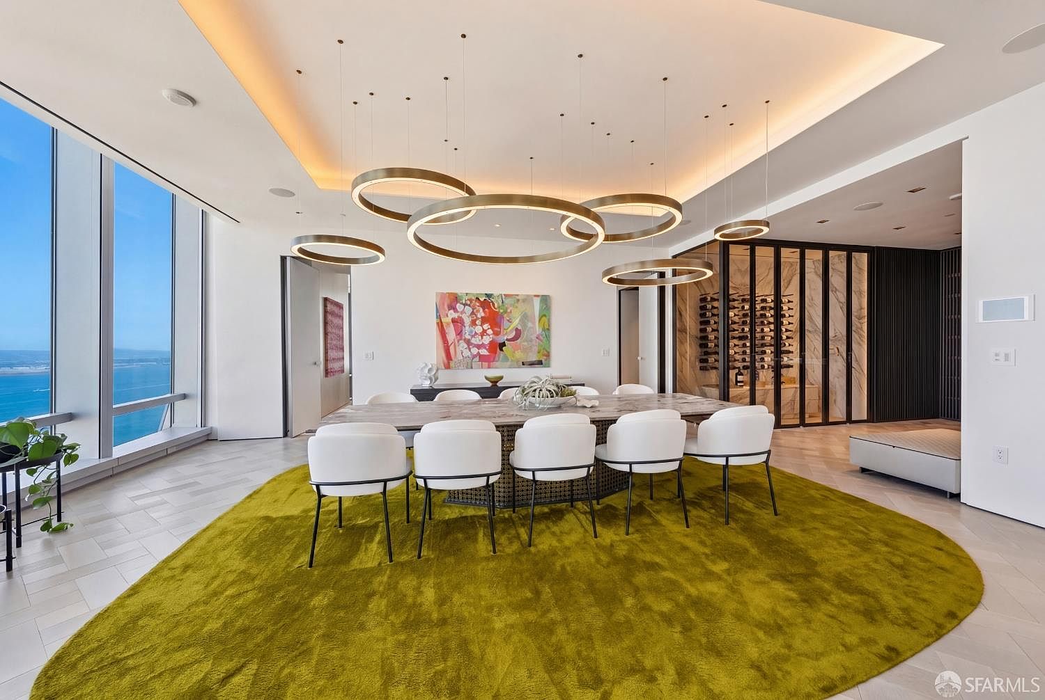 This sophisticated dining room features a large stone-topped table surrounded by modern white chairs, all set atop a vibrant, circular chartreuse rug. Above, a striking arrangement of circular pendant lights creates a focal point, while floor-to-ceiling windows offer expansive views of the water. To the side, a glass-enclosed wine display adds a luxurious touch to the open-concept space.