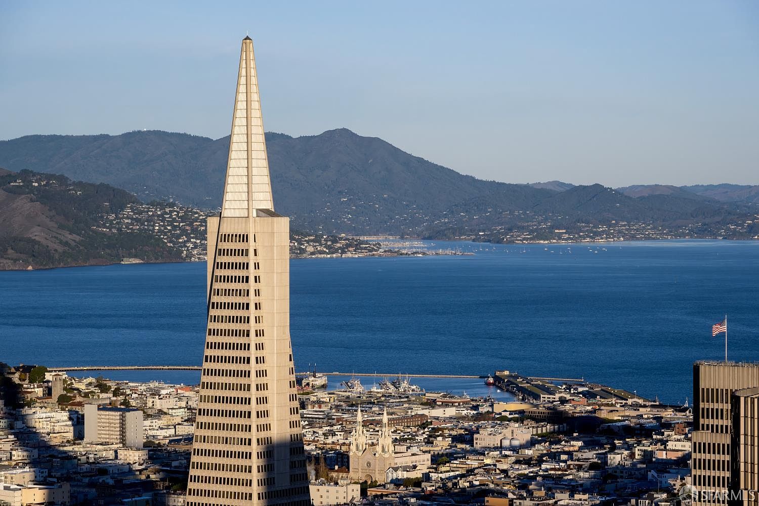 This high-angle aerial view captures the iconic Transamerica Pyramid in San Francisco, showcasing its distinctive tapered architectural design against the backdrop of the San Francisco Bay and distant mountains. The surrounding urban landscape features dense city blocks, historic buildings, and a clear view of the waterfront, emphasizing the property's prime location. The lighting suggests a clear, late afternoon setting, highlighting the building's geometric form and the expansive coastal scenery.