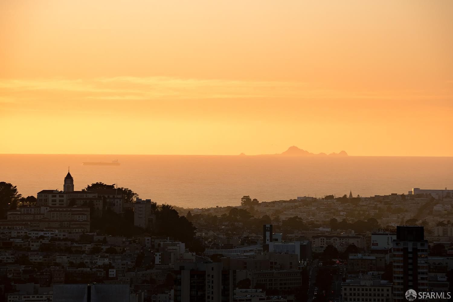 This wide-angle aerial view captures a stunning sunset over a dense urban landscape, likely in San Francisco, with the Pacific Ocean and distant islands visible on the horizon. The foreground features a mix of residential buildings and lush greenery, while a large cargo ship is silhouetted against the glowing orange sky. The perspective provides a cinematic sense of scale, highlighting the property's prime location and expansive coastal views.
