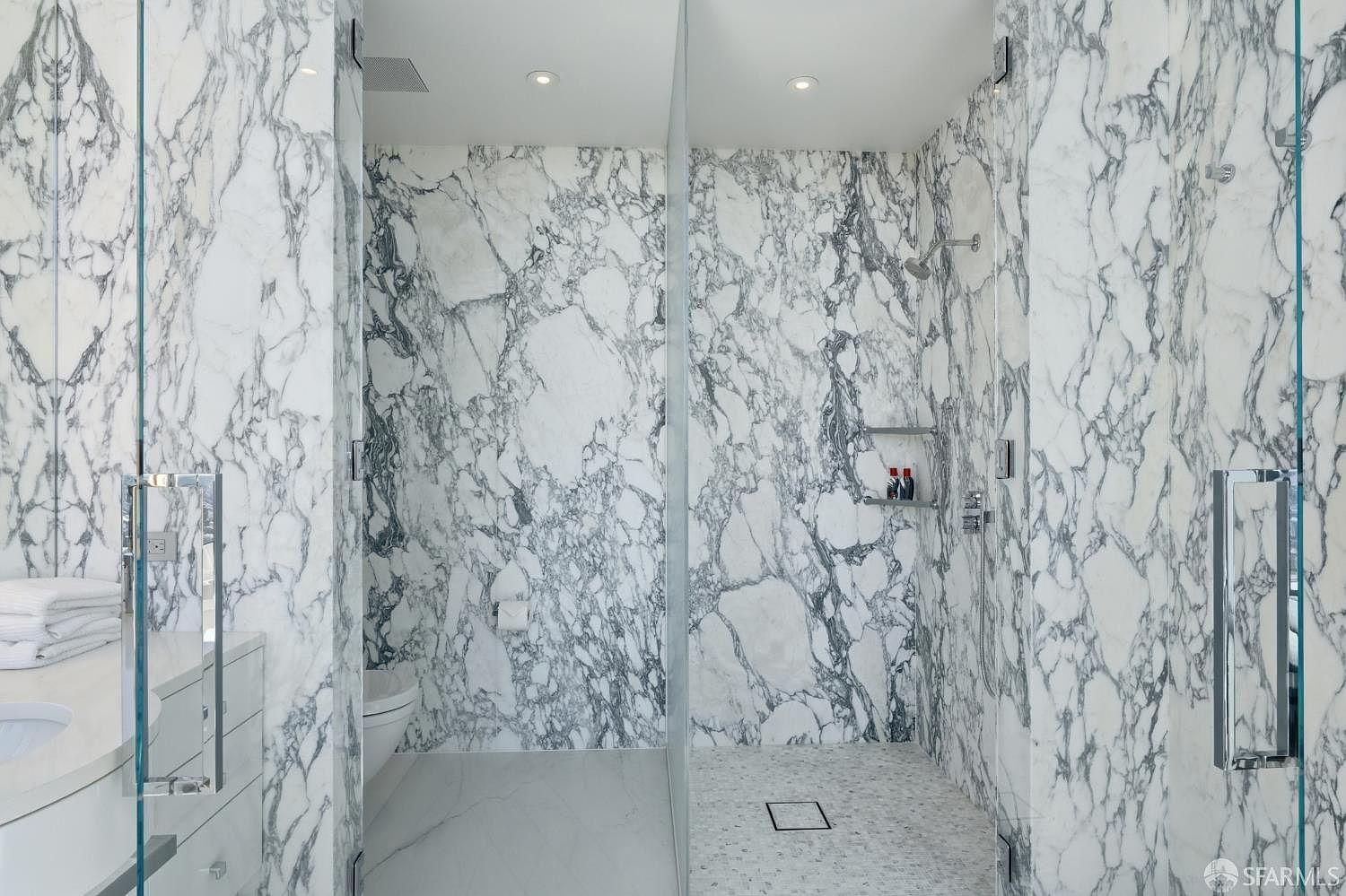 This luxurious bathroom features extensive use of patterned stone throughout the walls and shower enclosure, creating a cohesive and high-end aesthetic. The space includes a glass-enclosed walk-in shower with a rainfall showerhead and a separate toilet area, all accented by polished chrome fixtures. The bright, clean lighting and reflective surfaces contribute to a sophisticated and spa-like atmosphere.