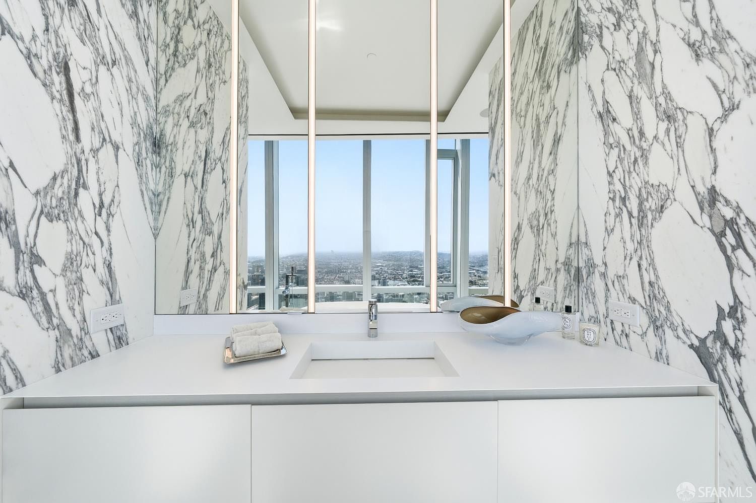 This modern bathroom features a sleek, minimalist vanity with a white countertop and a rectangular integrated sink. The space is framed by striking, high-contrast veined stone walls that extend behind a large mirror, which reflects a panoramic city view through the window. The composition is symmetrical and clean, emphasizing a luxurious, high-rise aesthetic.