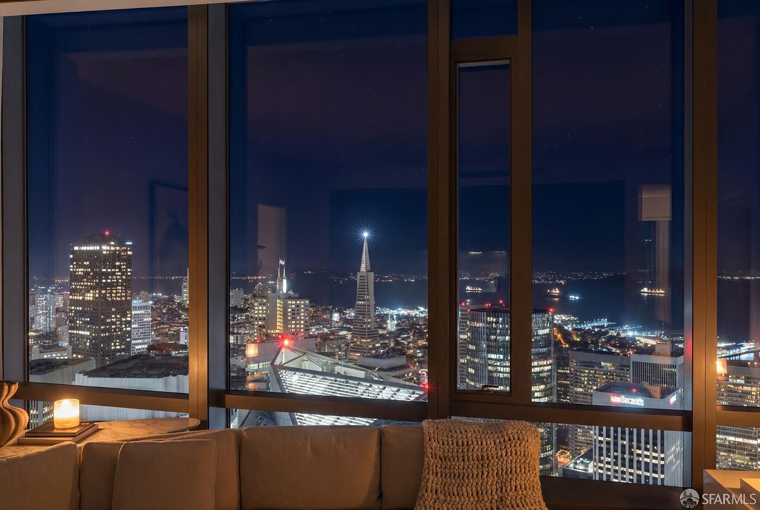 This interior view captures a sophisticated living room space framed by expansive floor-to-ceiling windows that offer a breathtaking, panoramic night view of the San Francisco skyline, including the iconic Transamerica Pyramid. The foreground features a plush, neutral-toned sofa topped with a textured throw blanket and a small side table holding a lit candle, creating a warm and inviting atmosphere. The composition emphasizes the contrast between the cozy, dimly lit interior and the vibrant, glittering urban landscape outside.