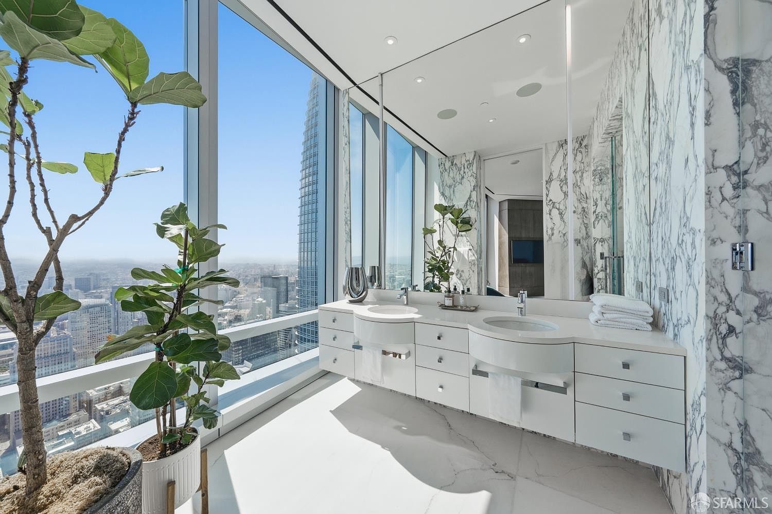 This luxurious primary bathroom features a sleek, double-vanity setup with white cabinetry and integrated sinks, set against a backdrop of striking, veined stone wall cladding. The space is defined by expansive floor-to-ceiling windows that provide a dramatic, unobstructed panoramic view of the city skyline. A large potted plant adds a touch of organic texture to the bright, minimalist aesthetic, creating a sophisticated and airy atmosphere.