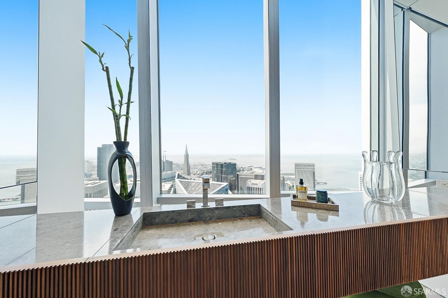 This high-rise bathroom features a sleek, stone-topped vanity with a recessed sink, positioned directly in front of expansive windows that offer a panoramic view of the city skyline and bay. The vanity is accented by a textured, slatted wood front, while a minimalist vase with bamboo and a small tray of toiletries add a refined touch. The perspective is eye-level, emphasizing the seamless integration of luxury interior design with the dramatic urban backdrop.