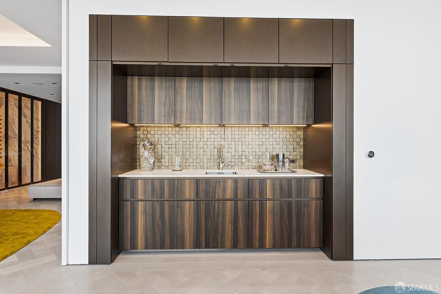 This image showcases a sleek, modern wet bar area integrated into a wall with dark, vertically grained wood cabinetry. The space features a light-toned stone countertop, a small undermount sink, and a textured, neutral-toned mosaic tile backsplash illuminated by under-cabinet lighting. The composition is centered and symmetrical, highlighting the sophisticated contrast between the dark cabinetry and the bright, minimalist surrounding walls.