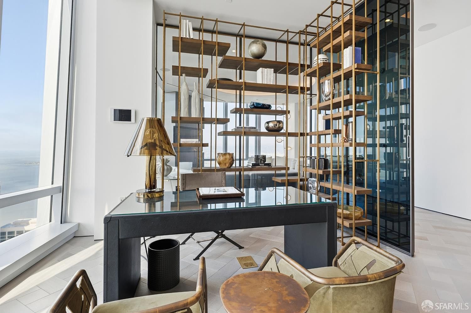 This sophisticated home office features a sleek glass-top desk and a modern, open-concept metal shelving unit that serves as a stylish room divider. The space is bathed in natural light from expansive floor-to-ceiling windows that offer a panoramic view of the water. With its contemporary furniture, neutral color palette, and high-rise perspective, the room exudes a professional and luxurious atmosphere.