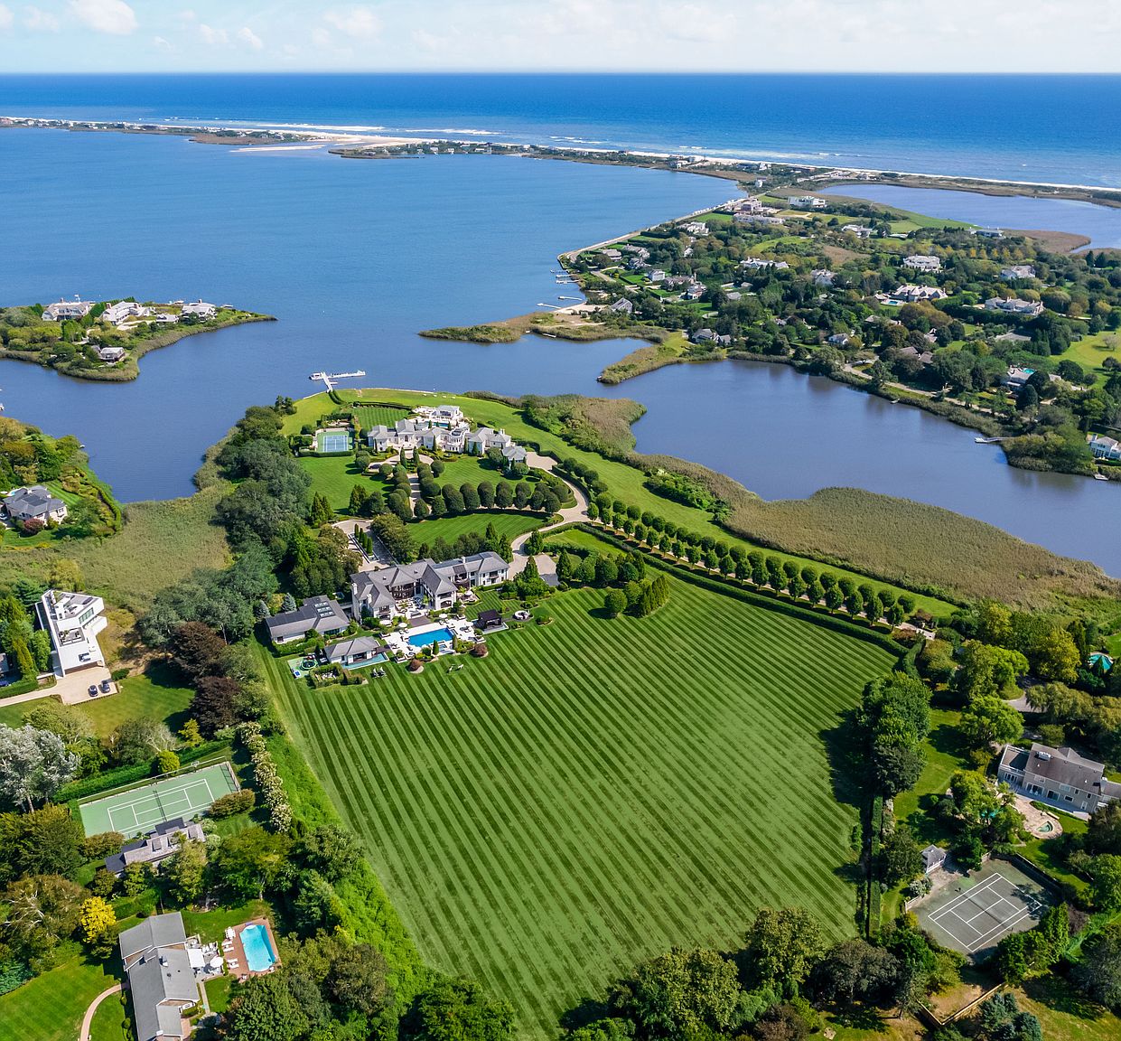 This aerial view showcases an expansive estate featuring a meticulously manicured lawn with striped patterns, a large main house with a pool, and several outbuildings. The property is surrounded by water on multiple sides, offering stunning waterfront views and a sense of seclusion. The overall impression is one of luxury and grandeur.