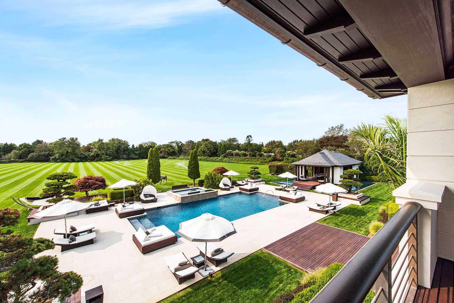 This image showcases a luxurious backyard featuring a large swimming pool and spa area, surrounded by meticulously manicured lawns and landscaping. Several lounge chairs and umbrellas are arranged around the pool, offering ample space for relaxation. A charming gazebo is visible in the background, adding to the property's appeal and creating a serene outdoor oasis.