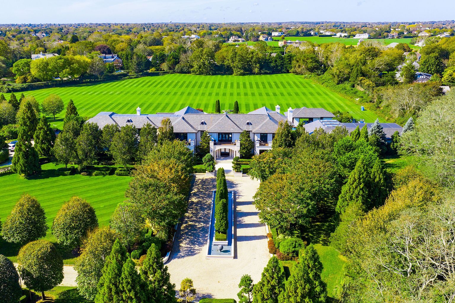 This aerial shot showcases a grand estate with meticulously manicured grounds. The property features a large, symmetrical lawn with striped patterns, a stately home with a gray roof, and a long, tree-lined driveway leading to the entrance. The overall impression is one of luxury and expansive space, perfect for highlighting the property's scale and landscaping.