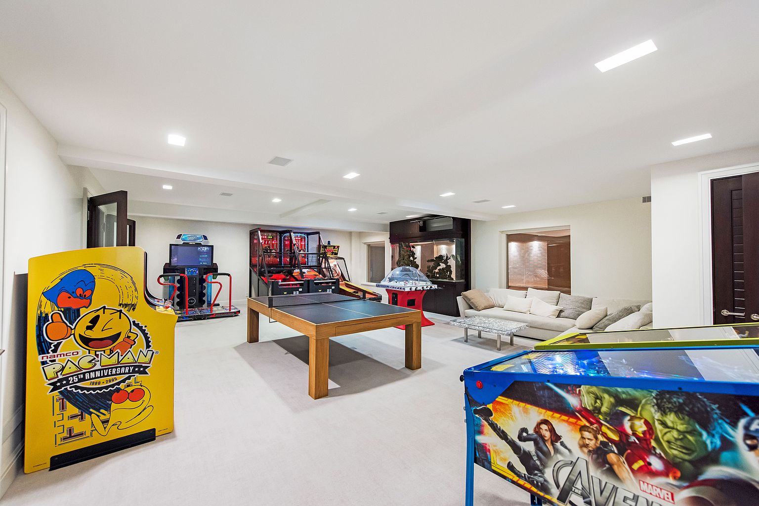 This is a spacious basement featuring a variety of entertainment options, including arcade games, a ping pong table, and a large fish tank. The room is well-lit with recessed lighting and has a neutral color palette, creating a versatile space for recreation and relaxation. A comfortable seating area with a sofa is also visible in the background.