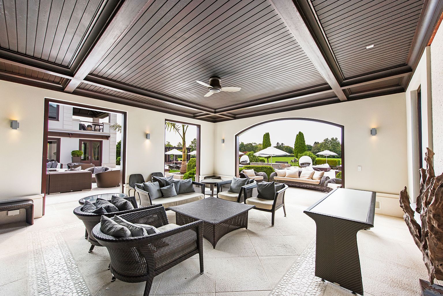 This image showcases a luxurious outdoor patio area, featuring elegant wicker furniture with plush cushions arranged around a woven coffee table. The space is covered by a sophisticated dark wood ceiling with recessed lighting and a ceiling fan, creating a comfortable and stylish lounge area. The patio opens to a beautifully landscaped garden with manicured hedges and sun umbrellas, offering a seamless transition between indoor and outdoor living.