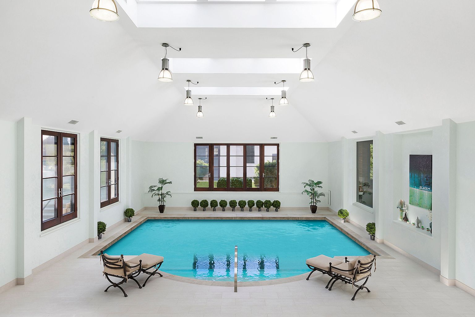 This is an interior shot of a luxurious indoor pool area. The pool is rectangular with turquoise water, surrounded by a light-colored tile deck. The room features multiple windows providing natural light, along with overhead lighting fixtures. Two lounge chairs are placed on either side of the pool, inviting relaxation.