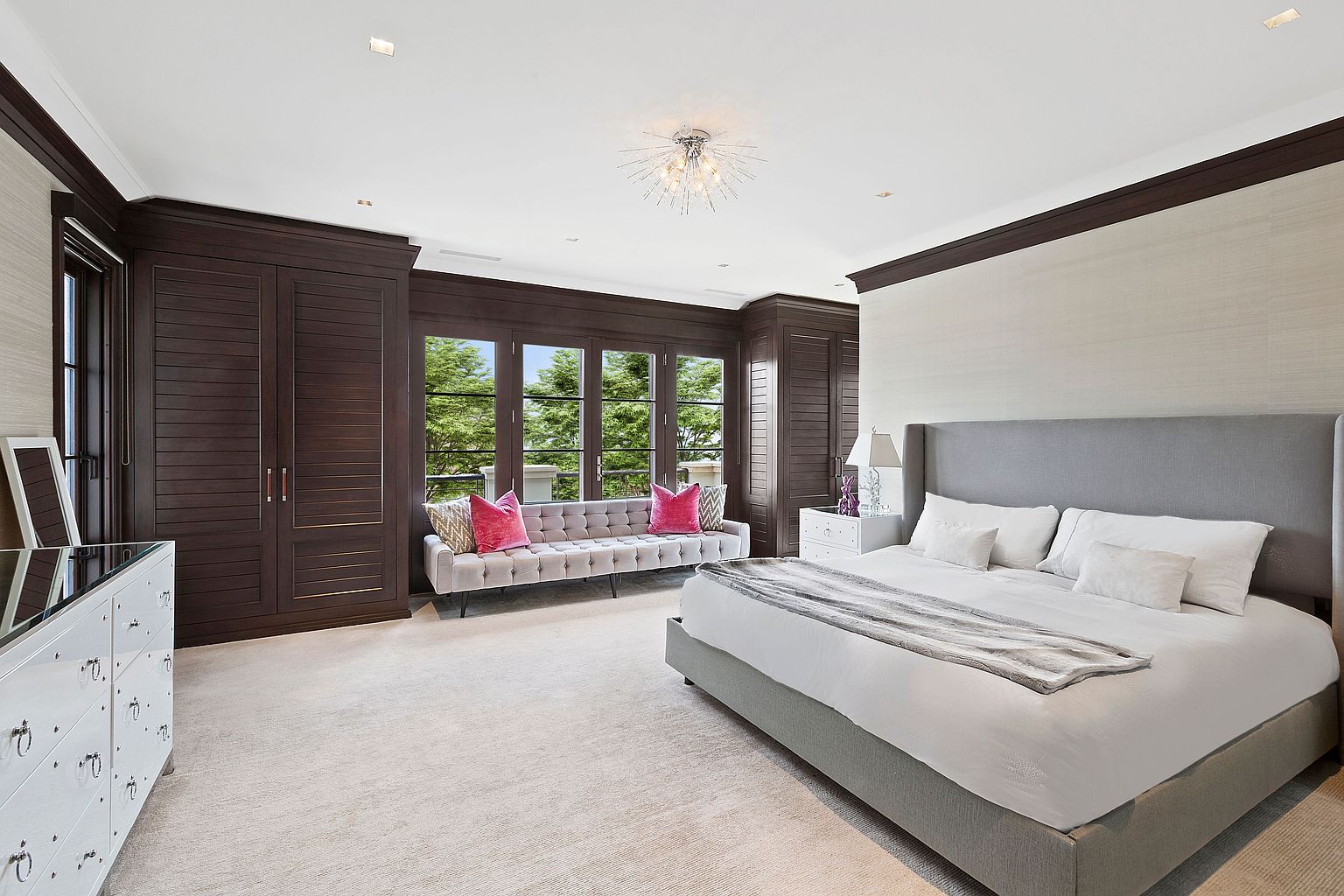 This is a luxurious primary bedroom featuring a large gray upholstered bed with white linens and a gray throw blanket. The room includes dark wood built-in wardrobes and a window seat with pink pillows, providing ample storage and a cozy seating area. The overall aesthetic is elegant and serene, with a neutral color palette and sophisticated lighting.