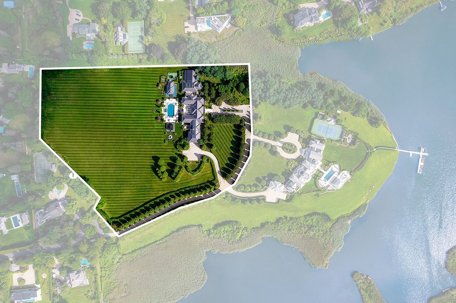 This aerial view showcases a sprawling estate with meticulously manicured lawns, a large swimming pool, and multiple buildings. The property is bordered by water on one side, offering stunning waterfront views. The overall impression is one of luxury and privacy.