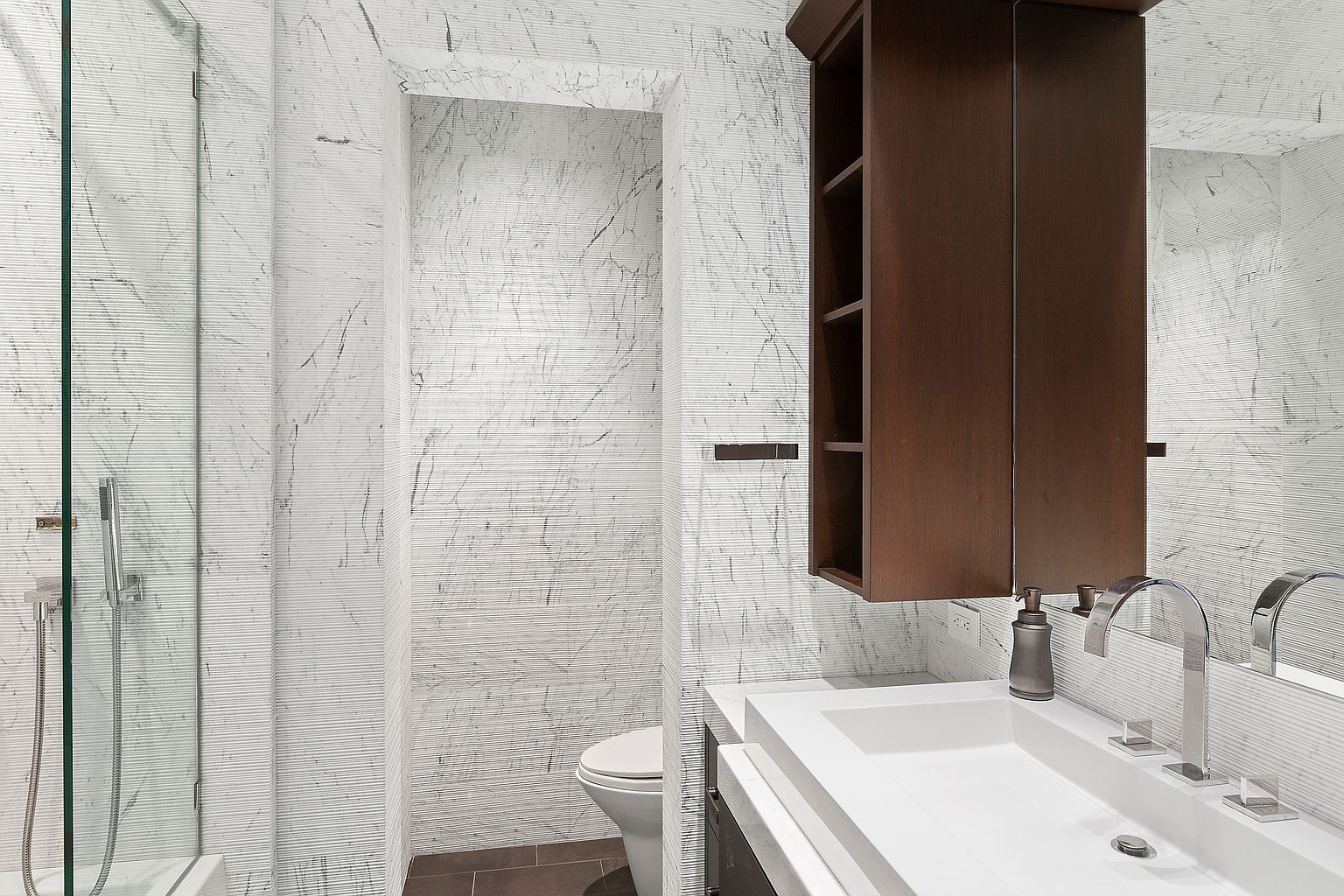 This is a modern bathroom featuring a white vanity with a vessel sink and a dark wood cabinet. The walls are covered in a textured white material with subtle gray veining, creating a clean and sophisticated look. A glass-enclosed shower is visible on the left, and a toilet is partially visible in the background, offering a glimpse into the layout of the space.