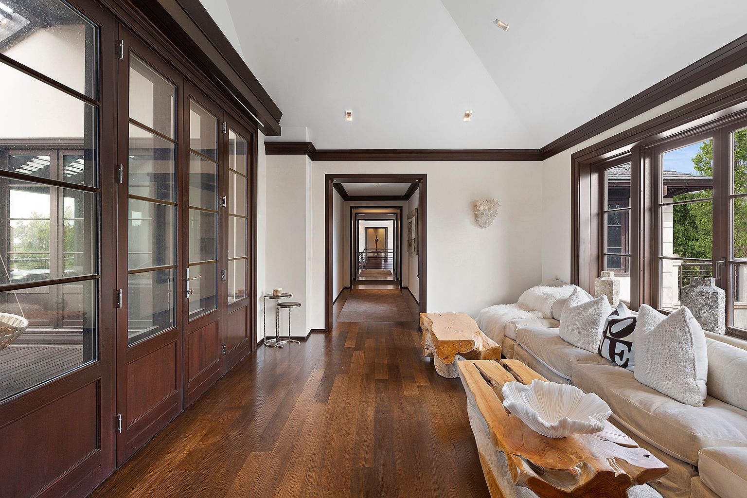 This interior shot showcases a long hallway with dark hardwood flooring and white walls, accented by dark wood trim. The hallway leads to a series of doorways, creating a sense of depth and spaciousness. To the right, a living area is visible with a light-colored sofa and unique wooden coffee tables, adding a touch of rustic elegance.