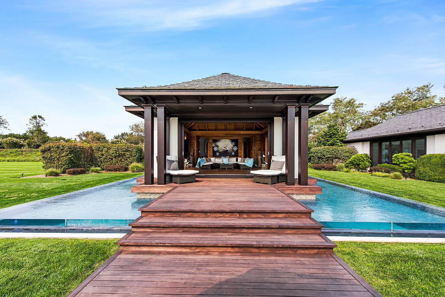 This image showcases a luxurious outdoor pool area featuring a modern pavilion with dark wood accents and a light-colored interior. The pavilion is accessed via a wooden walkway and steps that extend over the pool, creating a unique and inviting space. The surrounding landscape includes well-manicured lawns and mature trees, enhancing the property's appeal.