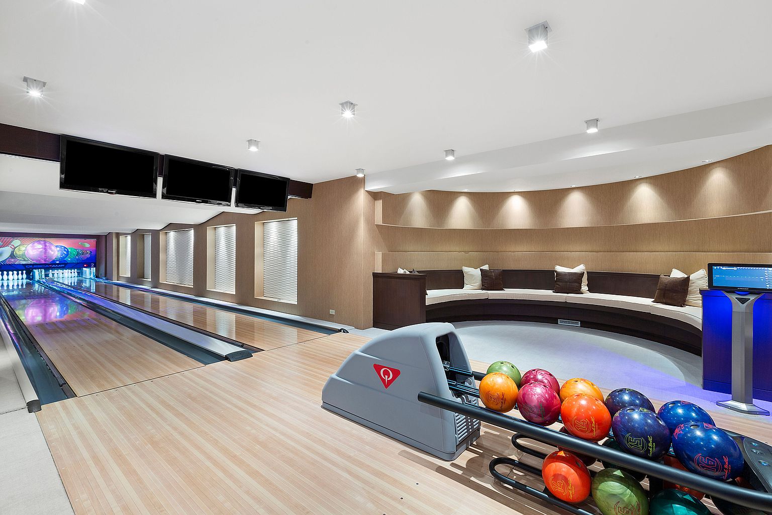 This is an interior shot of a basement bowling alley. The image showcases two bowling lanes with a ball return system and a seating area with a curved sofa. Three flat-screen televisions are mounted on the wall above the lanes, and the lighting is bright and modern.