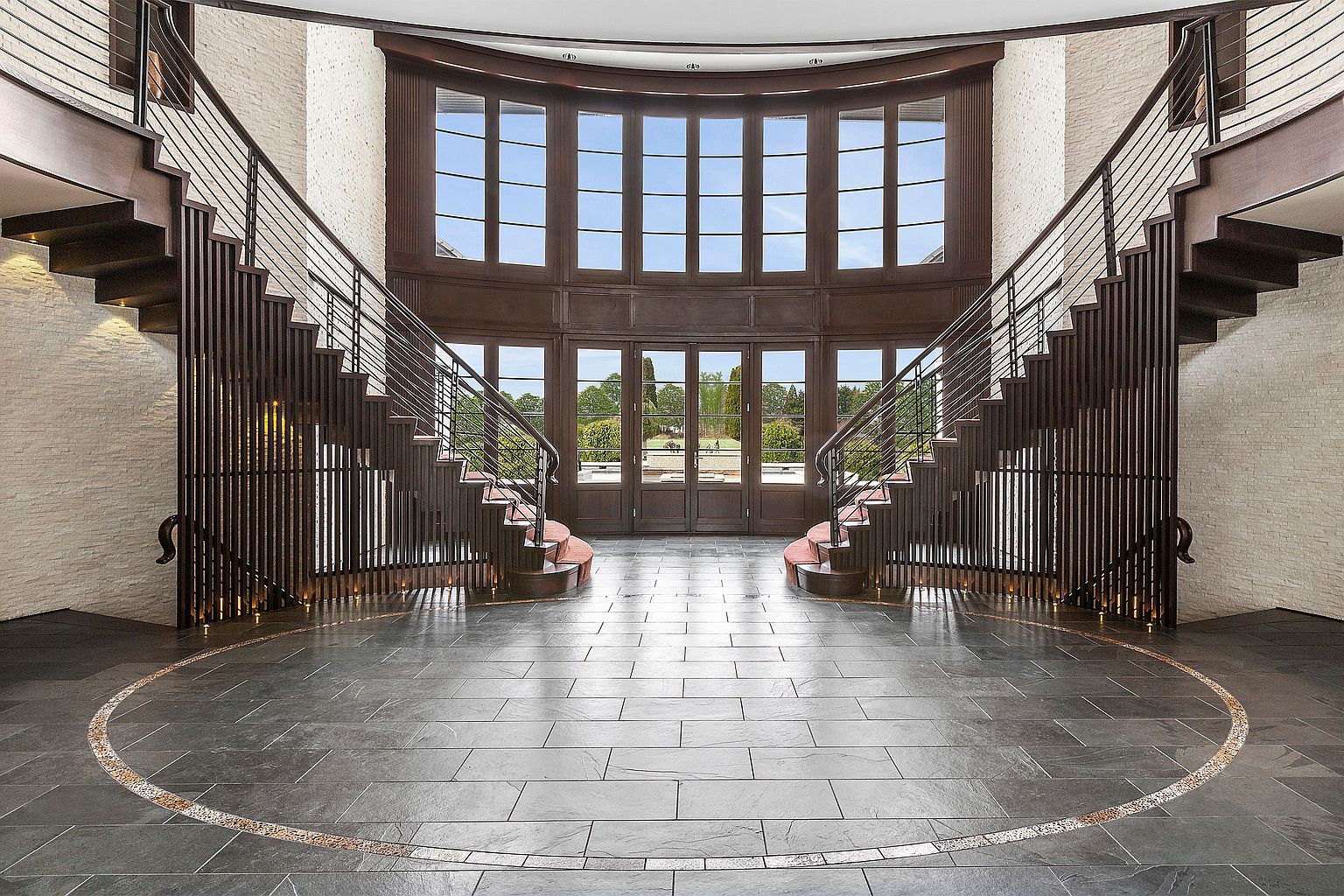 This grand foyer features dual staircases flanking a large window wall, providing ample natural light and a view of the exterior. The dark wood stairs contrast with the light-colored brick walls, and the slate tile flooring with a circular accent adds a touch of elegance. The overall impression is one of spaciousness and sophisticated design.