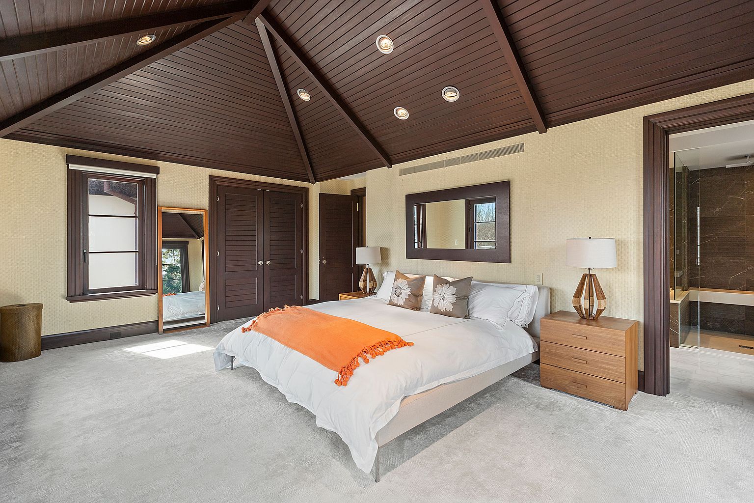 This is a primary bedroom featuring a king-size bed with white linens and an orange throw blanket. The room has a dark wood vaulted ceiling with recessed lighting, light yellow walls, and light gray carpet. There is a window, a full-length mirror, and a bathroom entrance visible.