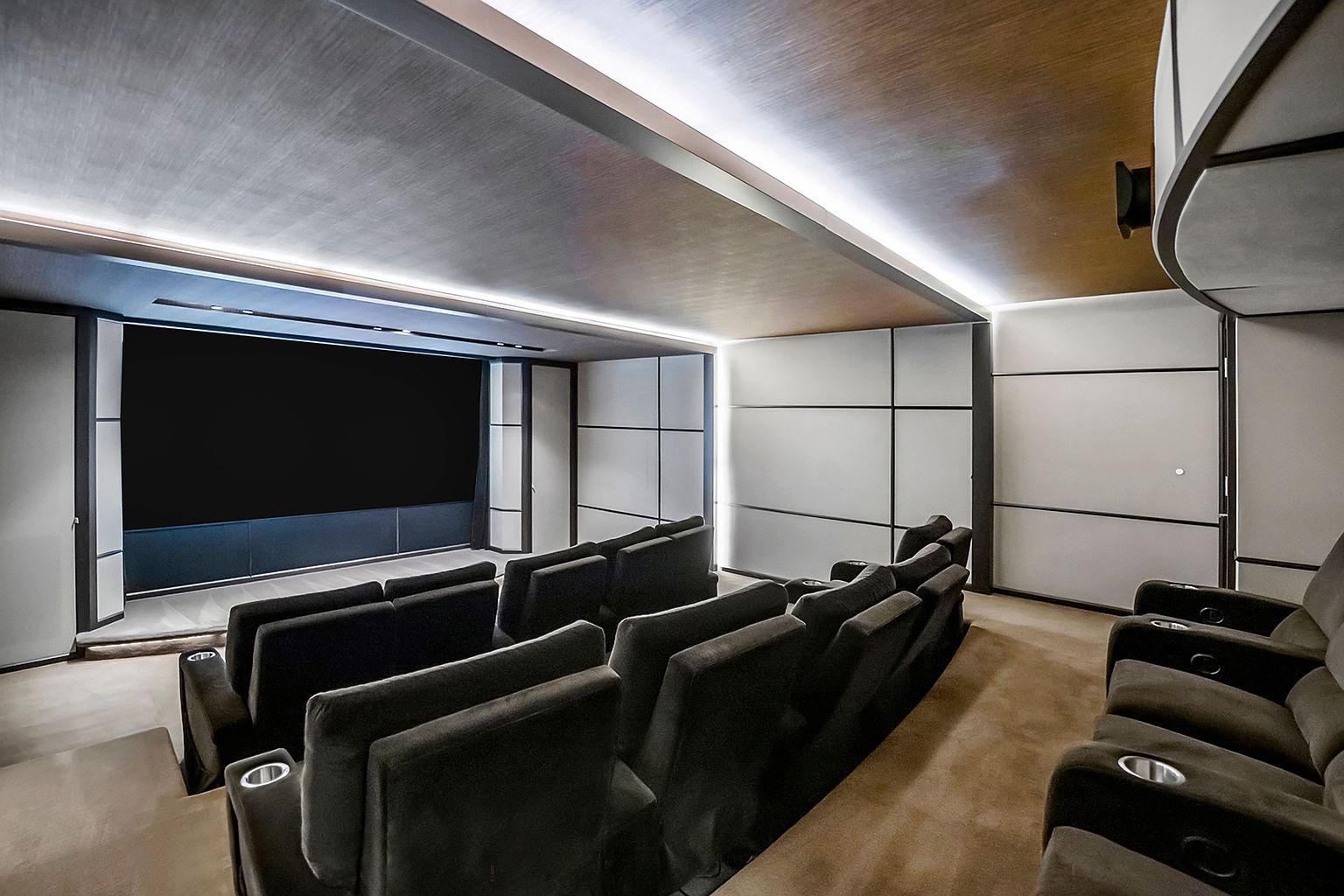 This is a home theater featuring multiple rows of plush, dark-colored seating facing a large screen. The walls are paneled with sound-dampening material, and the ceiling has recessed lighting, creating a cinematic atmosphere. The perspective is from the back of the room, looking towards the screen.