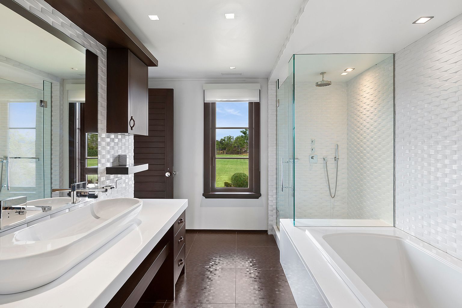 This is a well-lit primary bathroom featuring a modern design with a long, white countertop and dual vessel sinks. A large mirror and dark wood cabinetry add contrast, while a glass-enclosed shower and tub combination is visible on the right. The floor is tiled in a dark, mosaic pattern, and a window provides natural light and a view of the outdoors.