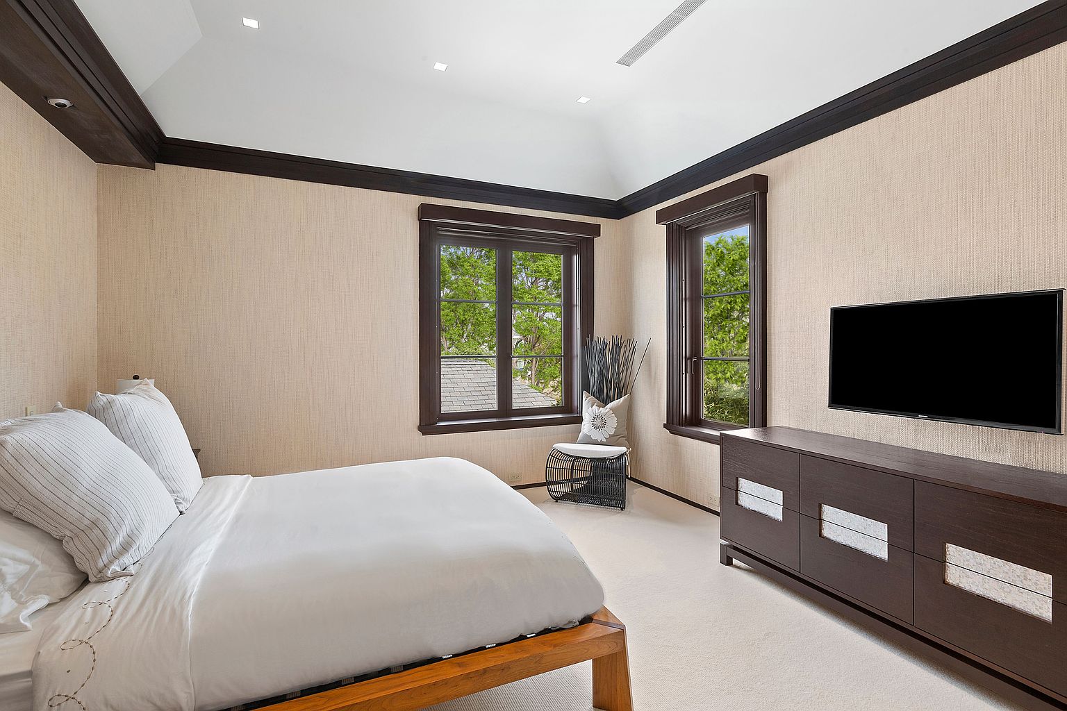 This is a well-lit primary bedroom featuring a king-size bed with white linens, a dark wood dresser with a flat-screen TV mounted above, and two windows providing natural light. The walls are covered in a textured wallpaper, and the room is trimmed with dark wood molding, creating a sophisticated and comfortable atmosphere. A small decorative chair sits near one of the windows.