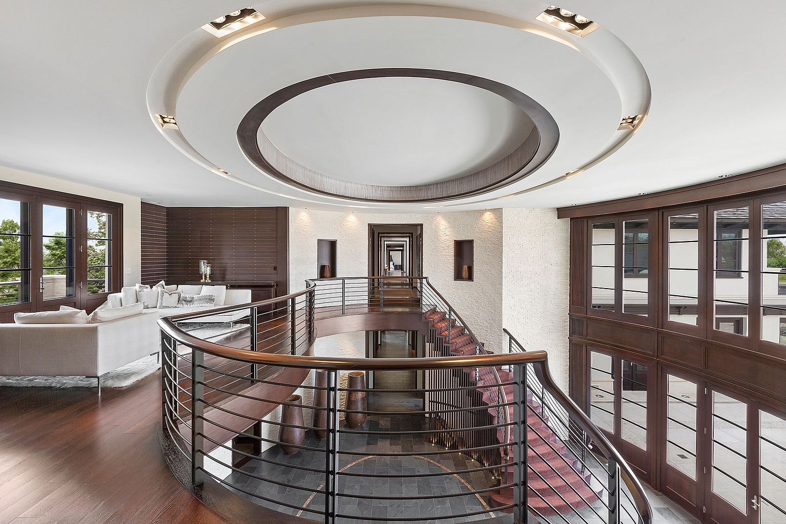 This is an interior shot of a luxurious home's hallway and staircase area. The space features a curved balcony with metal railings and wooden accents, overlooking a lower level with dark tiled flooring. The ceiling has a unique circular design with recessed lighting, and the walls are textured, creating an elegant and sophisticated atmosphere.